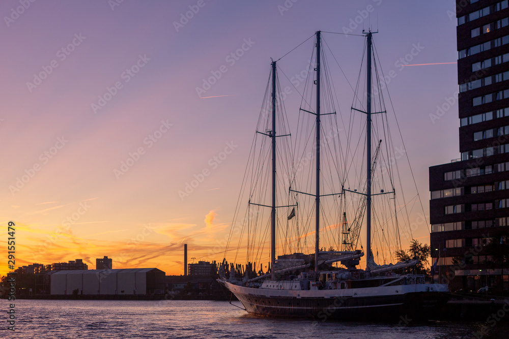 Docked traditional sailboat next to high rise building against a deep purple and orange sunset sky