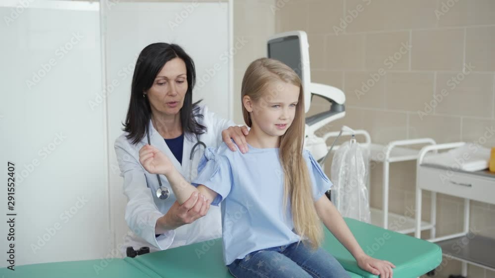 Tracking medium shot of female pediatrician in white coat raising arm of smiling little girl sitting on couch in doctors office and palpating her shoulder Tracking medium shot of female pediatrician in white coat raising arm of smiling little girl sitting on couch in doctors office and palpating her shoulder