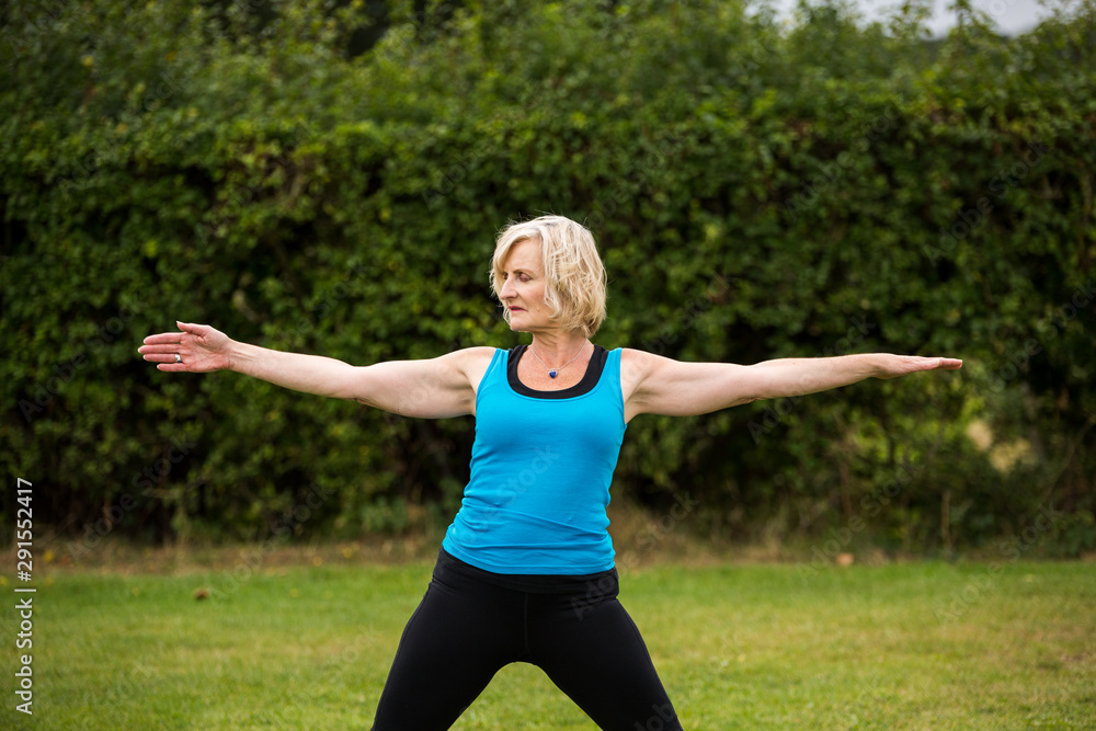 A middle aged woman practicing yoga barefoot outside in a grassy park. She is wearing a bright blue vest and black leggings. The style of yoga she is doing is Hatha Yoga A middle aged woman practicing yoga barefoot outside in a grassy park. She is wearing a bright blue vest and black leggings. The style of yoga she is doing is Hatha Yoga