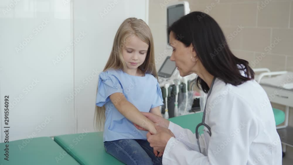 Tracking medium shot of female pediatrician in white coat talking friendly to little girl sitting on couch in doctors office and checking her arm Tracking medium shot of female pediatrician in white coat talking friendly to little girl sitting on couch in doctors office and checking her arm