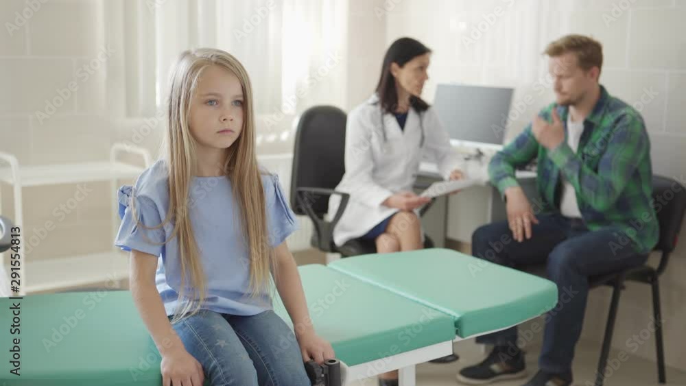 Pensive little girl sitting on couch in doctors office while female pediatrician explaining diagnosis to her frustrated father in background, tracking shot Pensive little girl sitting on couch in doctors office while female pediatrician explaining diagnosis to her frustrated father in background, tracking shot