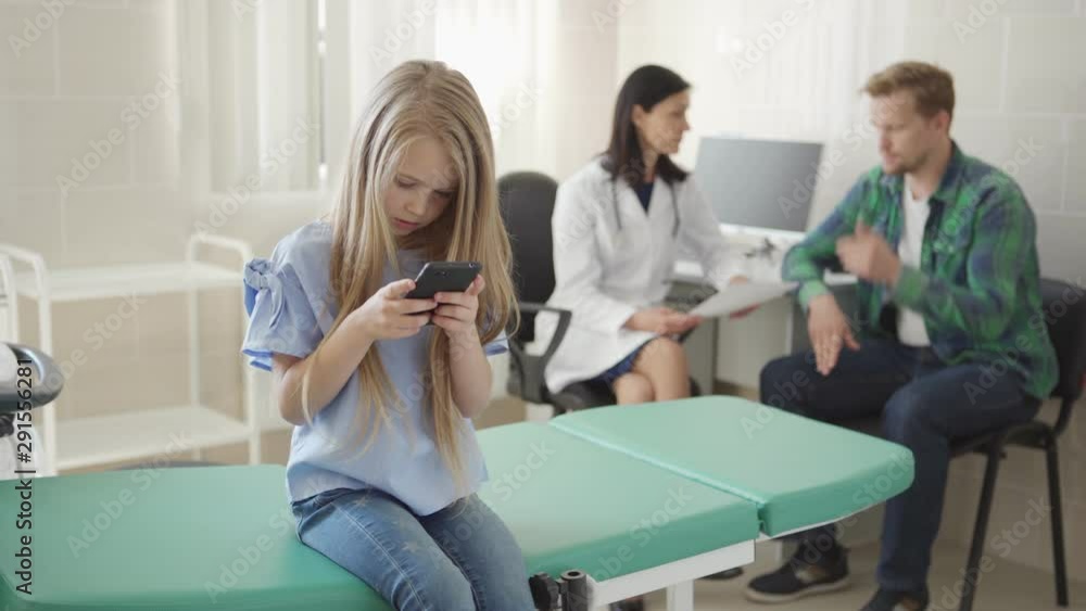 Tracking shot of little girl sitting on couch in doctors office and playing on cell phone while pediatrician telling bad news to her frustrated father in background Tracking shot of little girl sitting on couch in doctors office and playing on cell phone while pediatrician telling bad news to her frustrated father in background
