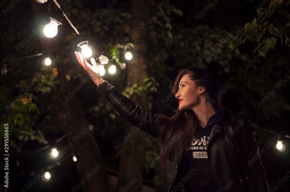 a beautiful woman in black stands under a garland of bright luminous bulbs and reaches out to them. Concept for a successful search for inspiration and ideas.