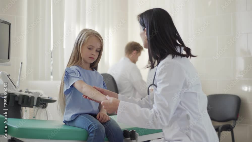 Tracking medium shot of female pediatrician in white coat talking friendly to little girl sitting on couch in doctors office while checking her arm, male doctor working at desk in background Tracking medium shot of female pediatrician in white coat talking friendly to little girl sitting on couch in doctors office while checking her arm, male doctor working at desk in background