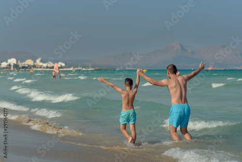 Cute European boy and his dad are running along beautiful seacoast. They are happy to spend their vacations together. Their hands are wide open. Back view.