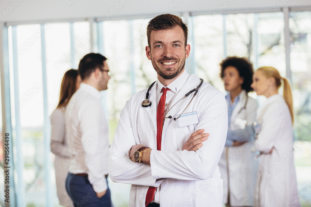 Male doctor with colleagues in background, doctor looking at camera with emergency team in background Male doctor with colleagues in background, doctor looking at camera with emergency team in background