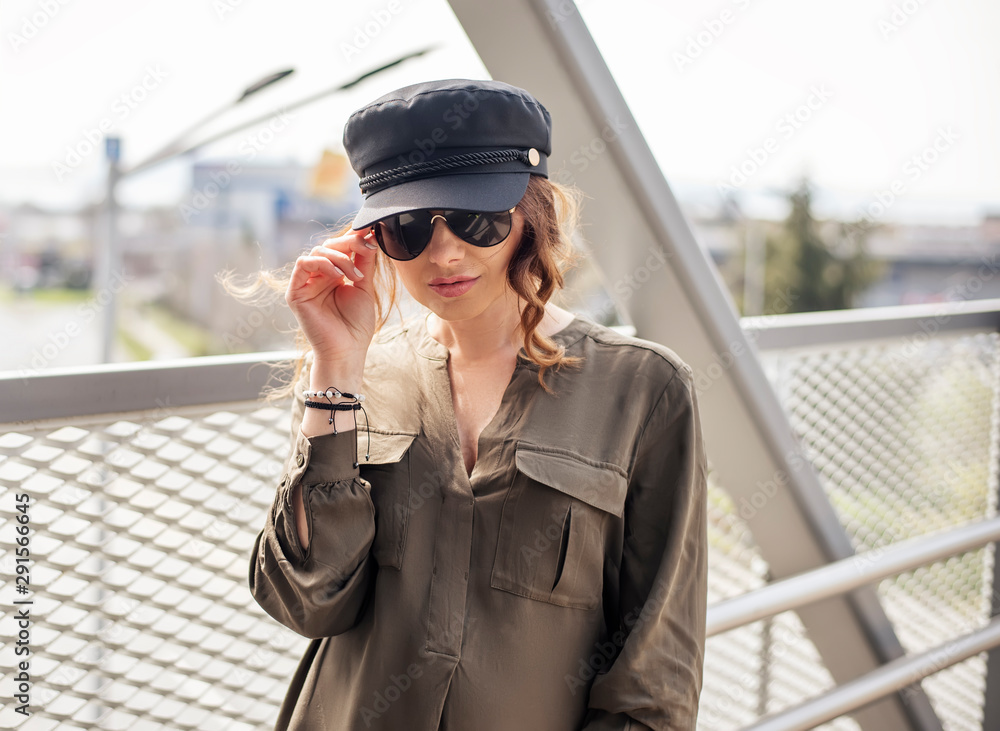 Portrait of fashion female model dressed casual in a grungy location. Fashion blogger in military shirt and hat on a pedestrian bridge