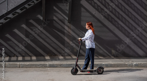 A red-haired girl in a white shirt drives an electric scooter along the wall. A business woman in a trouser suit and red high heels rides around the city in a modern car. Business woman on a scooter.