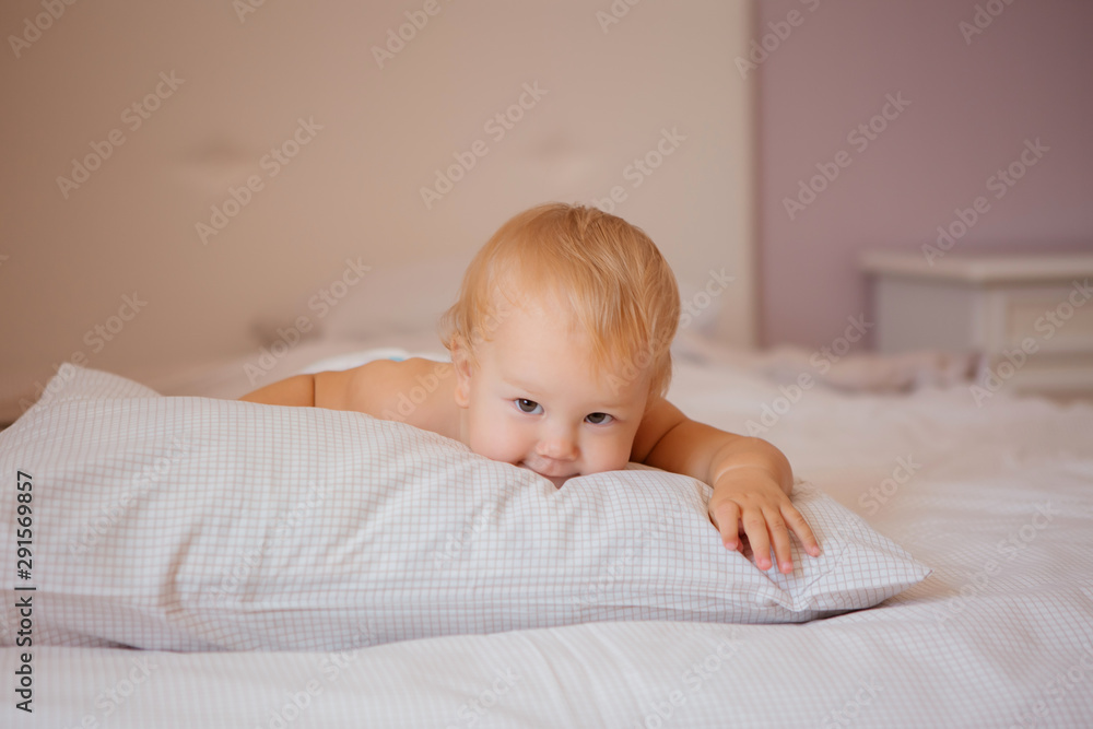 blonde baby in a diaper is lying on a bed covered with a white sheet