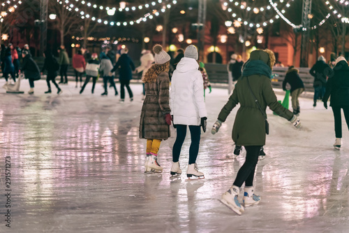 Group of girs skating back to us. Girlfriends ice skating in city park, snowy evening. Healthy outdoor winter activity