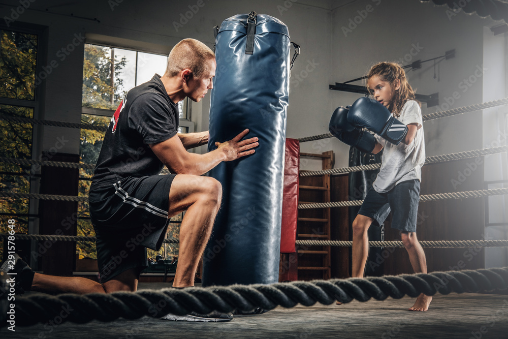 Experienced boxer trainer is training new little girl boxer for special competitions using punching bag.