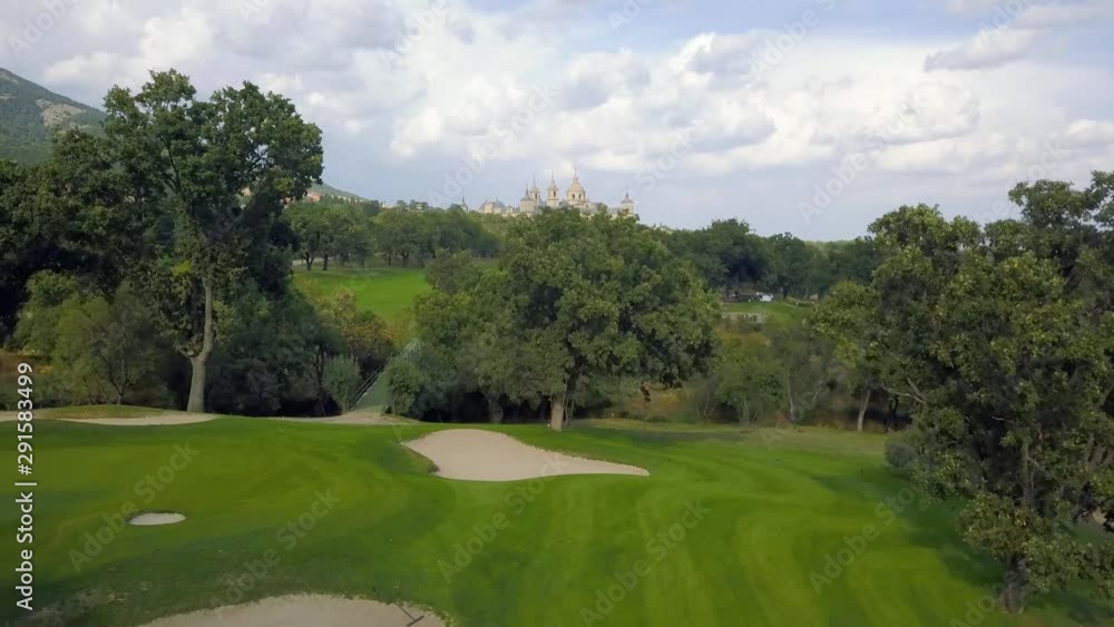 golf course in a forest of ash with the historic monastery of escorial crowning a hill, aerial view 