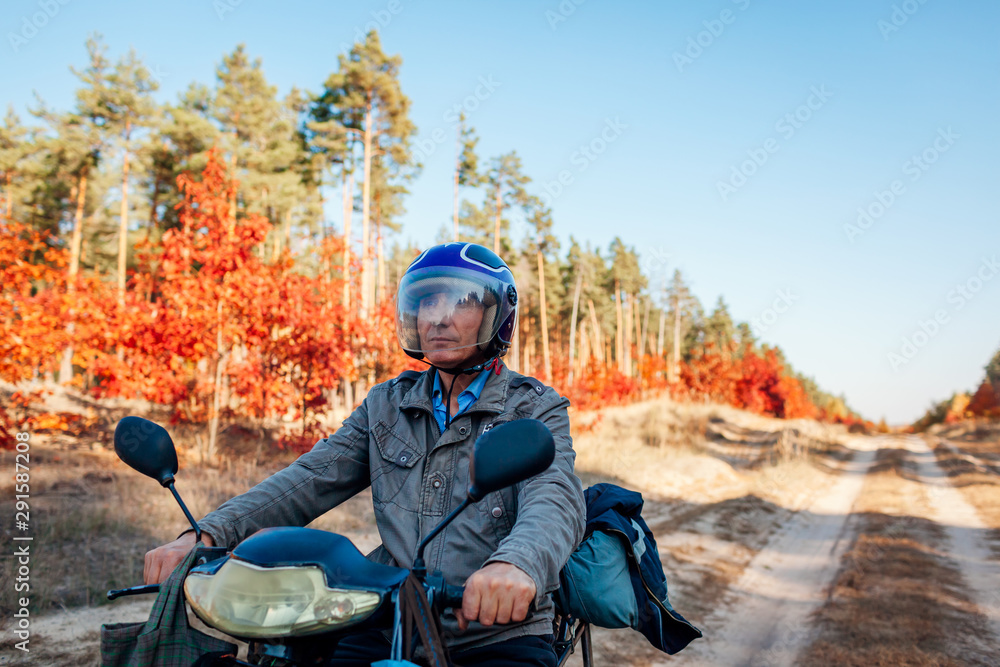 Senior man riding scooter on autumn forest road. Driver in helmet riding moped