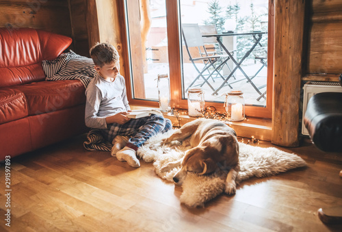 Boy reading book on the floor near slipping his beagle dog on sheepskin in cozy home atmosphere. Peaceful moments of cozy home concept image.