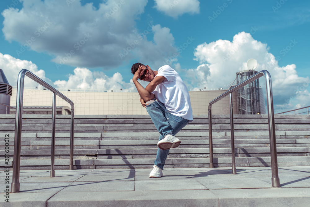 young guy a dancer in a white T-shirt, jeans, dancing break dance in summer in the city, in sunglasses, white sneakers background building clouds, active hip hop, youth lifestyle