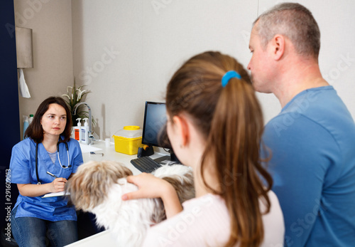 Professional woman veterinarian consulting anxious father and daughter with small dog in clinic