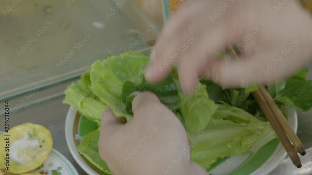 A daylight extreme closeup shot of a customer wrapping a mini rice pancake in a leaf of green lettuce, dipping it in a chili vinegar sauce and then taking a bite out of it. A daylight extreme closeup shot of a customer wrapping a mini rice pancake in a leaf of green lettuce, dipping it in a chili vinegar sauce and then taking a bite out of it.