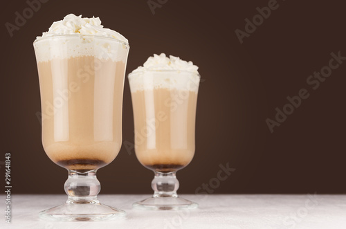 Two hot coffee drinks with whipped cream in wineglass in dark brown interior on white wooden table, copy space.