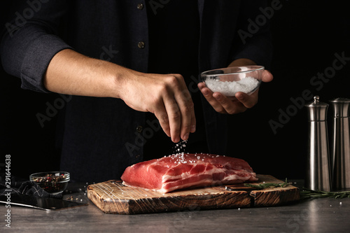 Fotografie Man salting fresh raw meat on table against dark background, closeup