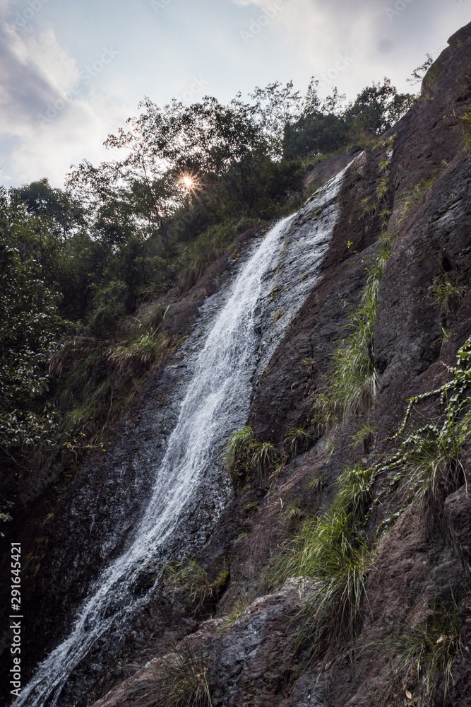tall waterfall rushing down the cliff under cloudy sky with rock surface filled with green plants and sunlight shine behind foliage and heavy cloud
