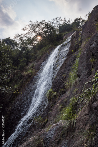 tall waterfall rushing down the cliff under cloudy sky with rock surface filled with green plants and sunlight shine behind foliage and heavy cloud