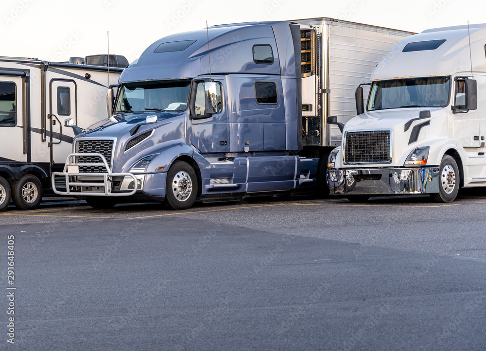 Gray and white big rig semi trucks with semi trailers standing in row on truck stop parking lot