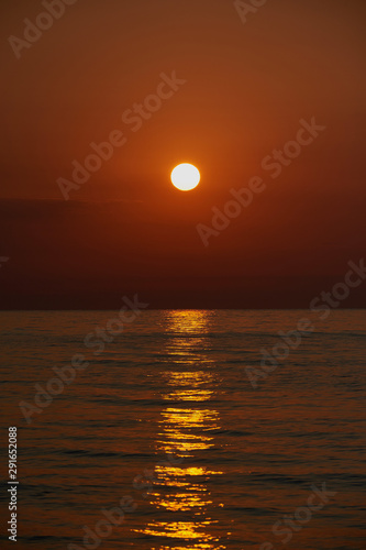Romantic sunset by a stony seashore. The sun sets over the horizon. The sun beams reflecting in the calm sea waters. Stony shore is washed by the gentle waves. The sky is turning yellow and orange.
