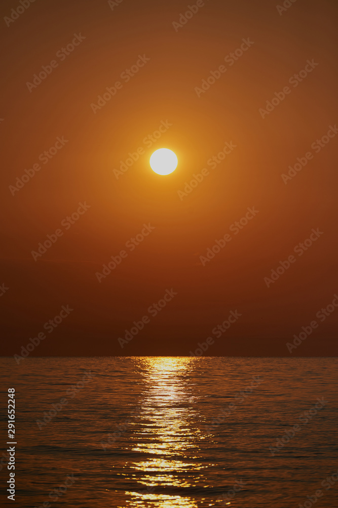 Romantic sunset by a stony seashore. The sun sets over the horizon. The sun beams reflecting in the calm sea waters. Stony shore is washed by the gentle waves. The sky is turning yellow and orange.