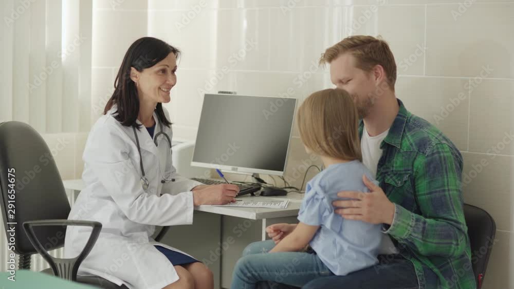 Middle aged female doctor in white coat sitting at desk in office and smiling while talking to father stroking gently back of little daughter sitting on his lap Middle aged female doctor in white coat sitting at desk in office and smiling while talking to father stroking gently back of little daughter sitting on his lap