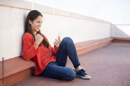 Chica adolescente celebrando buenas noticias on line sentada en la azotea photo