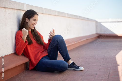 Chica adolescente celebrando buenas noticias on line sentada en la azotea photo