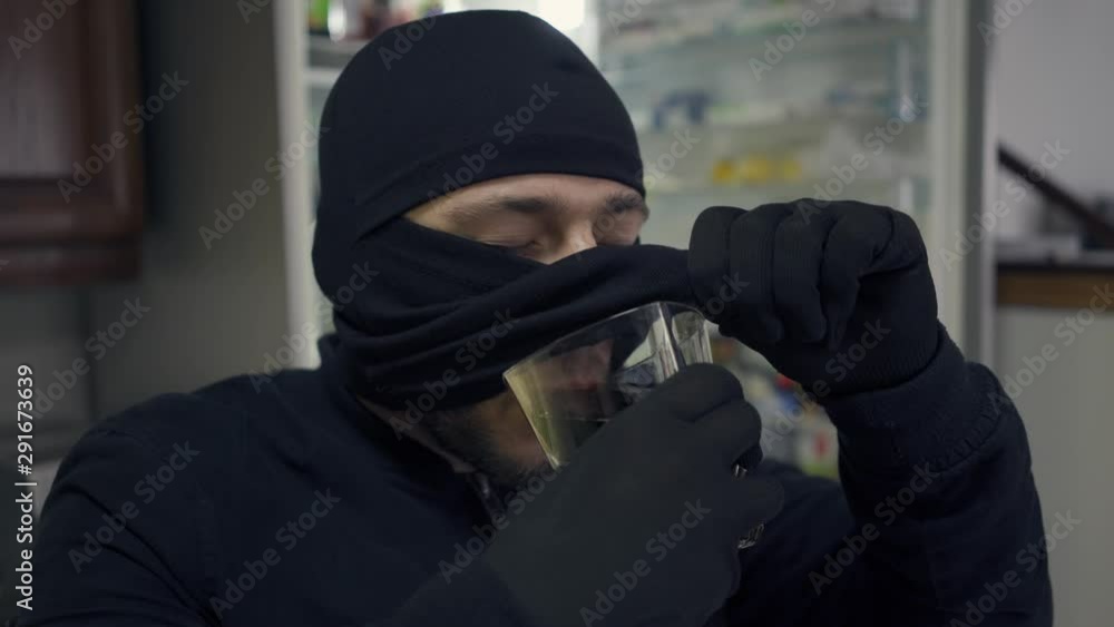 Close-up of young caucasian thief in black clothes raising his balaclava and drinking from the glass. The guy stealing things in rich house. Concept of offense against the law. Close-up of young caucasian thief in black clothes raising his balaclava and drinking from the glass. The guy stealing things in rich house. Concept of offense against the law.