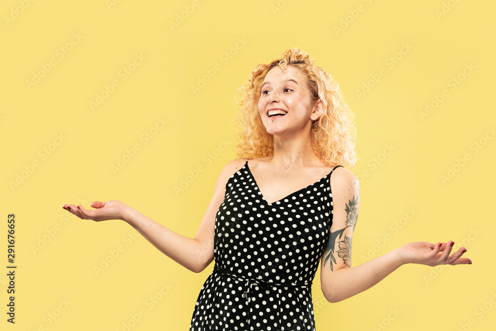 Caucasian young woman's half-length portrait on yellow studio background. Beautiful female model in black dress. Concept of human emotions, facial expression. Showing and inviting somebody. Caucasian young woman's half-length portrait on yellow studio background. Beautiful female model in black dress. Concept of human emotions, facial expression. Showing and inviting somebody.
