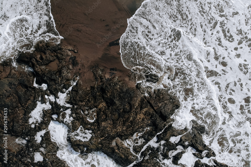 Fototapeta premium Top view aerial photo from flying drone of ocean landscape with big waves and rocks on black sand beach. Copy space for your advertising text message or promotional content.Perfect website background