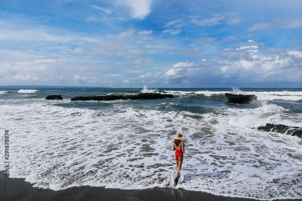 Aerial view of back view woman in red bikini and straw hat enjoing ocean with rocks and beach with black sand . Vacation in Bali. Photo from drone