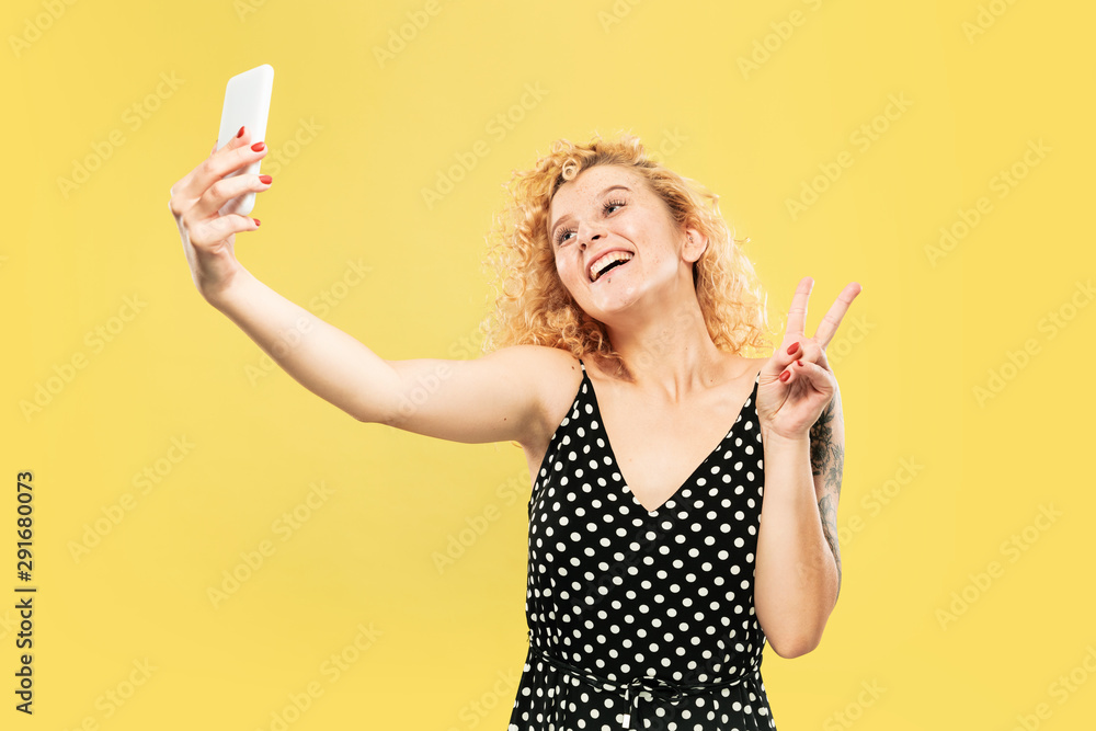 Caucasian young woman's half-length portrait on yellow studio background. Beautiful female model in black dress. Concept of human emotions, facial expression. Taking selfie or her own vlog, smiling.