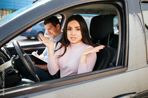 Male auto instructor takes exam in young woman. She sit on driver's place and look on camera with confusion. Stressed woman shake hands. Instructor sit behind and look down. © estradaanton