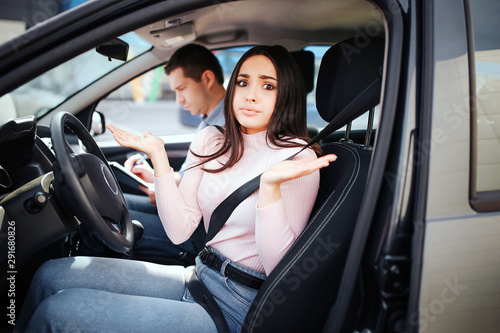 Male auto instructor takes exam in young woman. Sitting together in car. Confused woman look on camera and wave with hands. Busy instructor write. Exam time. © estradaanton