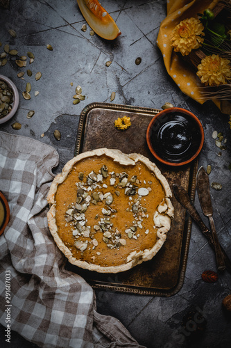pumpkin pie decorated with pumpkin seeds on metal tray with textile, fresh pumpkin slices, seeds and fall leaves