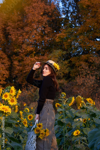  Beautiful lovely girl in a straw hat holding a wicker bag with flowers. Enjoying nature on a sunny field of sunflowers. Autumn time. The concept of happiness, tranquility, love of life.