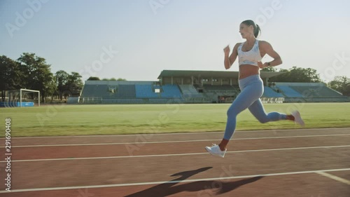 Beautiful Fitness Woman in Light Blue Athletic Top and Leggings Jogging in a Stadium. She is Running on a Warm Summer Afternoon. Athlete Doing Her Routine Sports Practice on a Track. Slow Motion. photo