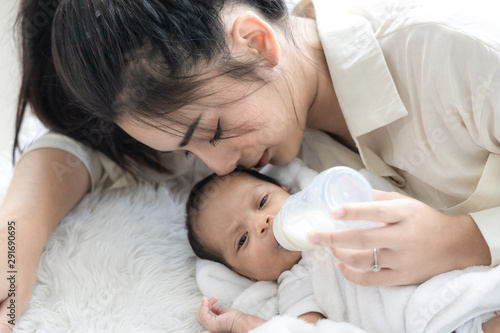 Young Asian mother hugging a cute newborn baby and feeding milk form bottle in white bedroom. Mother and child concept