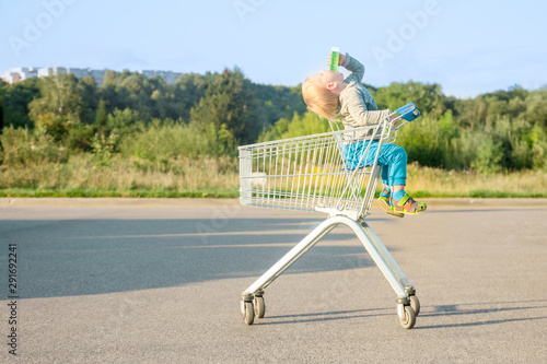 child boy blond in blue pants sits in a metal grocery trolley from a supermarket drinks juice throwing his head on an asphalt surface in the summer against a background of green plants photo