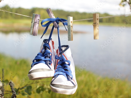 Pictures from a country holiday concept. Children's white sneakers with blue laces and colorful clothespins hanging on a clothesline. Blurred background of the lake and forest. Place for text.