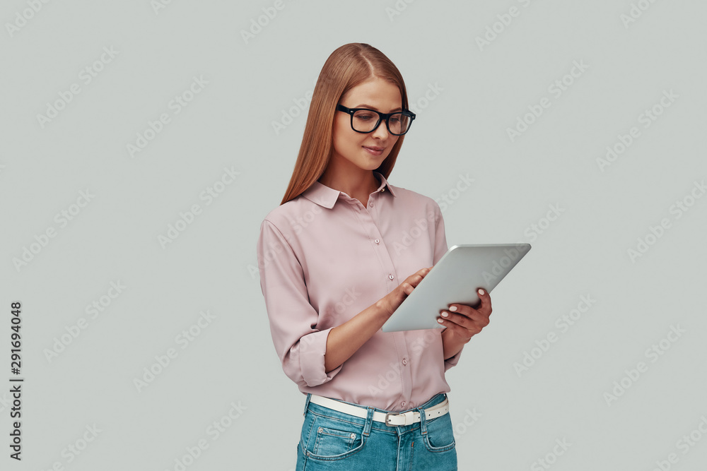 Attractive young woman in eyewear using digital tablet and smiling while standing against grey background Attractive young woman in eyewear using digital tablet and smiling while standing against grey background
