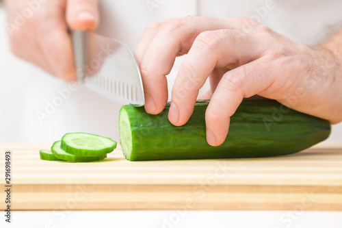 Man's hands cutting green cucumber with big knife on wooden board. Preparing vegetable ingredient for salad. Closeup. Front view.