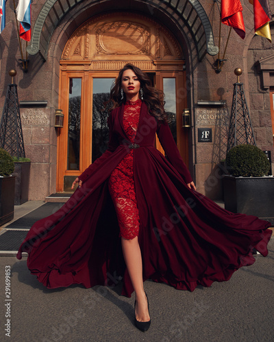 Young beautiful stunning woman in red luxury evening dress with flying hem walking city street on a sunny evening. Elegant lady with makeup and wavy brunette hair. Full length fashion portrait