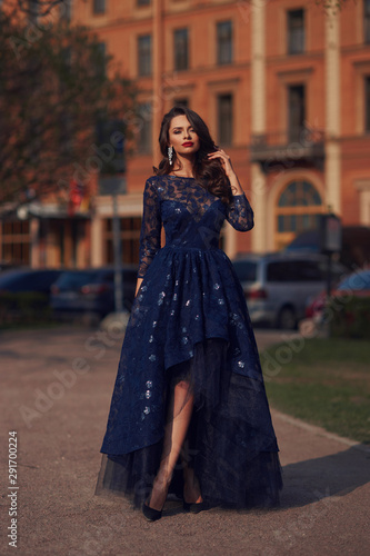 Elegant woman in blue ball gown dress standing and posing on a sunny evening at city street. Fashion model full length portrait