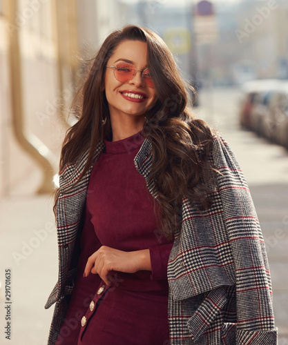Outdoor sunny portrait of young elegant stylish woman in burgundy color dress, gray coat and sunglasses. Caucasin pretty girl with makeup and wavy brown hair walking city street.