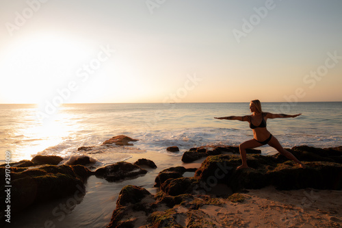 Young woman practicing yoga, standing in Virabhadrasana pose on the beach. Sunset time. Tegal Wangi beach, Bali